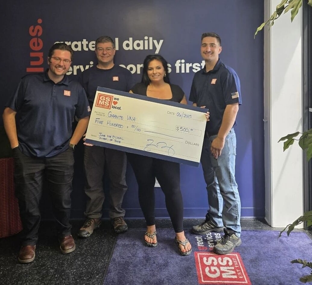 Four people from GSMS standing together inside an office, holding a large donation check for $500 made out to Granite VNA, dated April 2025. A wall behind them displays part of the company’s values, and they are smiling in support of the community initiative.