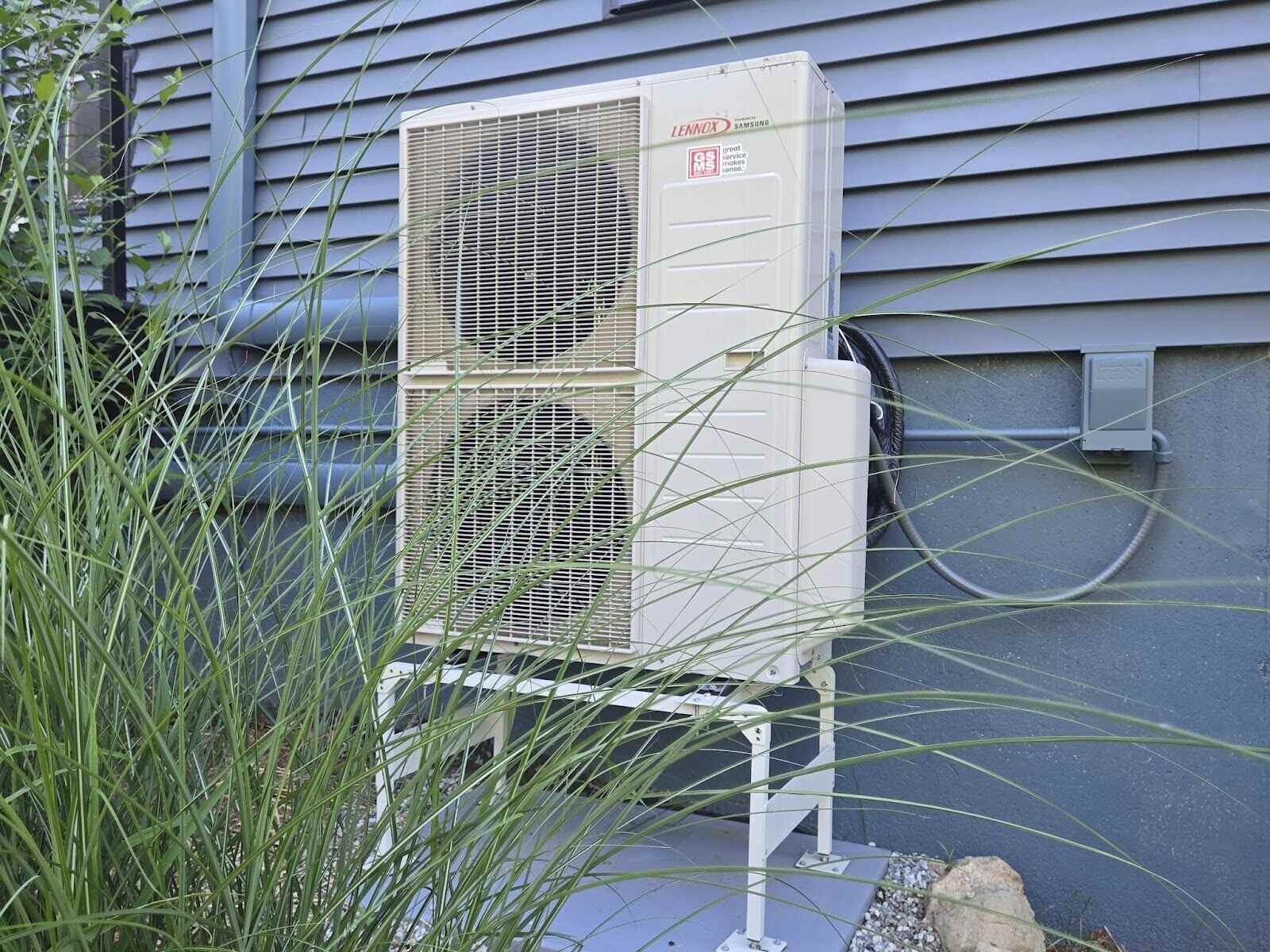Outdoor ductless mini split heat pump unit installed on a raised stand outside a home in Southern New Hampshire, partially hidden by tall ornamental grass.